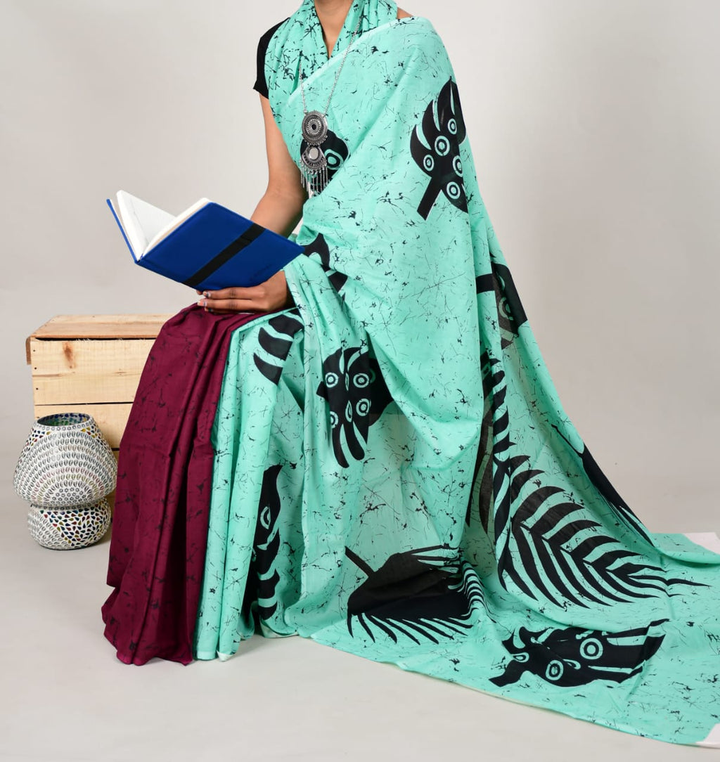Person wearing a light blue saree with black leaf patterns, sitting on a wooden box with a white background.